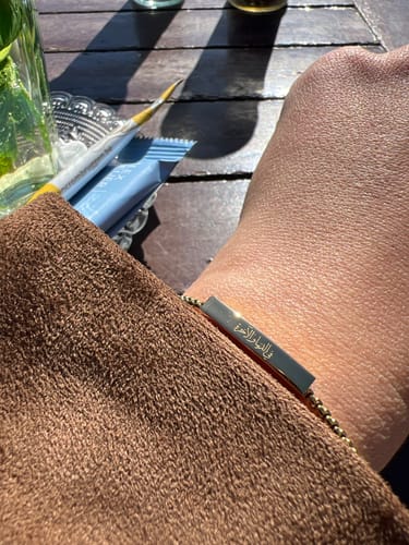 A reviewer's wrist wearing the 'In This World and the Next Bracelet', showing the detailed Arabic engraving on the bar.
