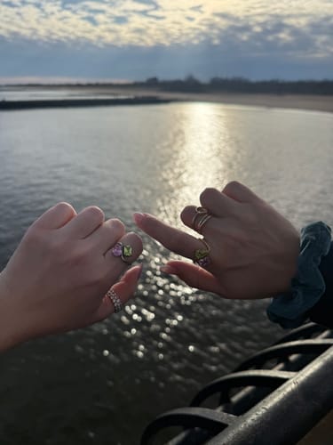 Two customers wearing matching Navae Birthstone Rings with purple and green stones, making a pinky promise gesture by the water.