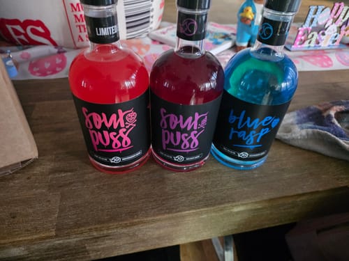 Reviewer's bottle of Sour Puss Grape liqueur sits alongside two other flavors on a wooden table.