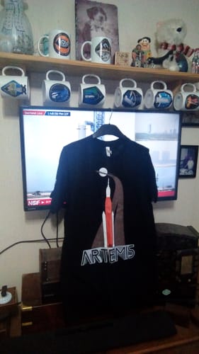 Customer's black Artemis II Vintage T-shirt with a rocket graphic displayed on a hanger in a room.