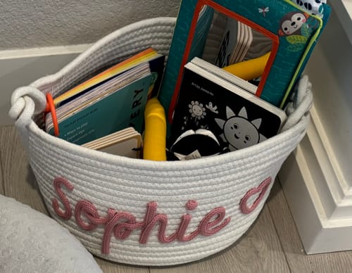 Customer's white personalized knitted basket with 'Sophie' and a heart icon in pink yarn, filled with books and toys.