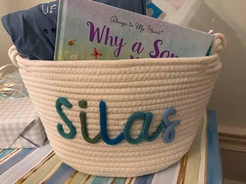 Customer's personalized cream knitted basket with the name 'Silas' in multicolor yarn, holding a book and other baby items.
