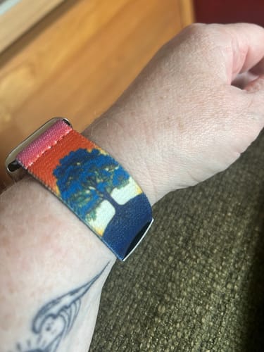 Customer's wrist showing a colorful watch band with a large blue tree design against a sunset-colored sky.
