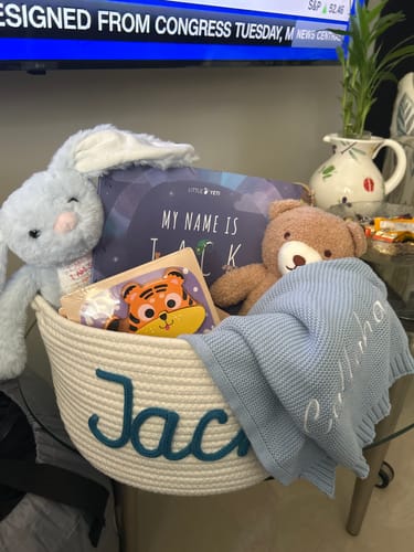 Reviewer's brown teddy bear Animal Plushie Toy sitting in a custom baby gift basket with other items.