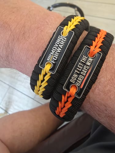 A close-up of a reviewer's wrist showing two Hard Days Paracord Bracelets, one black with yellow lacing and one with orange lacing.