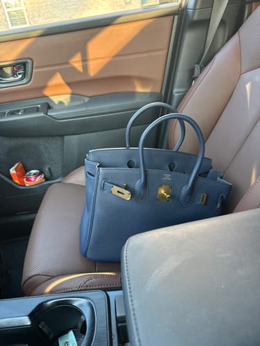 Customer's navy HM Birkin bag with gold hardware resting on the passenger seat of a car.