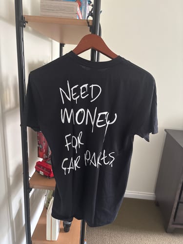 Customer's black 80Eighty® N$4Carparts Shirt on a hanger showing the white text "Need Money For Car Parts" on the back.