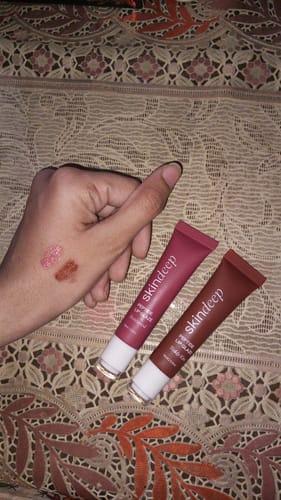 Reviewer holding two Peptide Lip Glaze tubes in Teddy Kiss and Pink Rose with swatches on their hand.