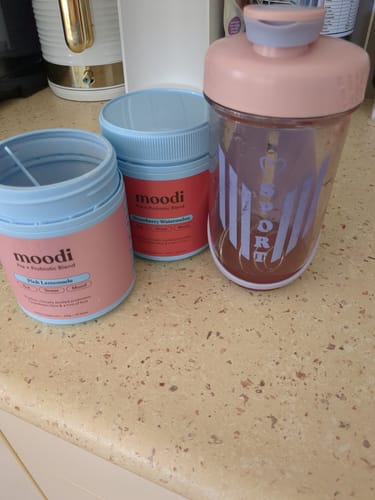 Customer's Pre + Probiotic Bundle tubs in Pink Lemonade and Strawberry Watermelon on a kitchen counter with a filled shaker cup.