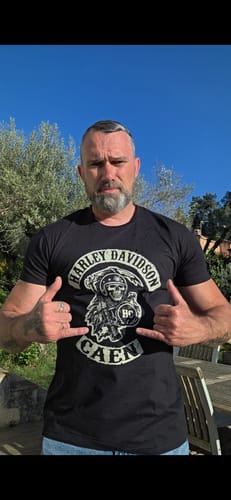 Client portant le t-shirt noir Faucheuse HC en extérieur, pointant le logo sur sa poitrine avec ses mains.