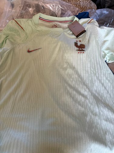 Un client montre le Maillot 2026 France Extérieur vert pâle, posé à plat avec son étiquette et son écusson.