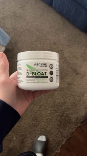 A customer holding a white tub of D-BLOAT powder in their hand, with a carpeted floor in the background.