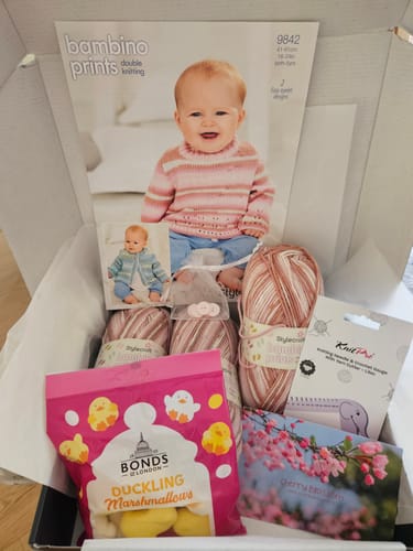 Reviewer's open Baby Girl Every Month box showing pink yarn, a baby knitting pattern, marshmallows, and accessories.