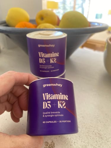 Main d'un client tenant un tube de Vitamine D3 + K2 avec un deuxième tube en carton en arrière-plan.