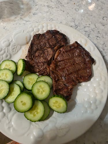 Customer's two cooked AAA Ribeye Steaks on a white plate with a side of sliced cucumbers.