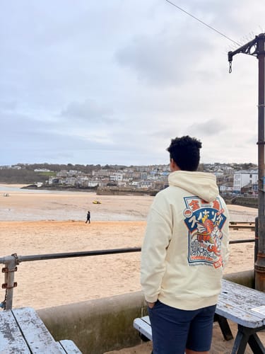 Reviewer from behind wearing the cream Koi King Hoodie, with the colorful back graphic visible against a beach backdrop.