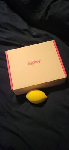 Reviewer's yellow Lem Clitoral Massager next to its beige and pink packaging on a dark bedspread.
