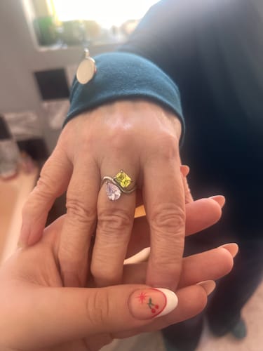 A person's hand wearing the Navae Birthstone Ring with a pear-cut purple stone and a radiant-cut yellow stone, holding another hand.