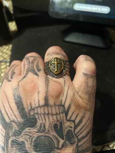 Close-up of a reviewer's tattooed hand wearing the Crafted Cross Ring, showing the detailed gold shield emblem with a cross.