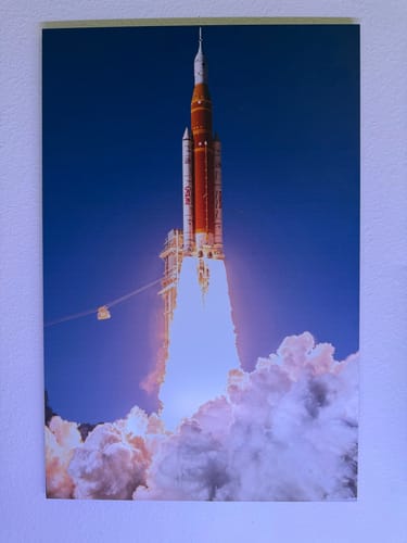 Reviewer's 'For All Humanity' print of a rocket launching, displayed on a light-colored wall.