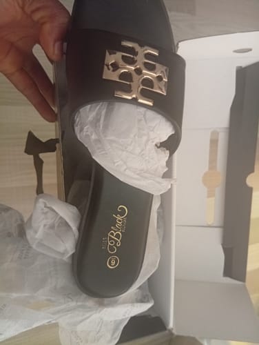 Close-up of a reviewer holding the black Miss Black Naledi 1 Sandal, showing its gold emblem, over the open box.