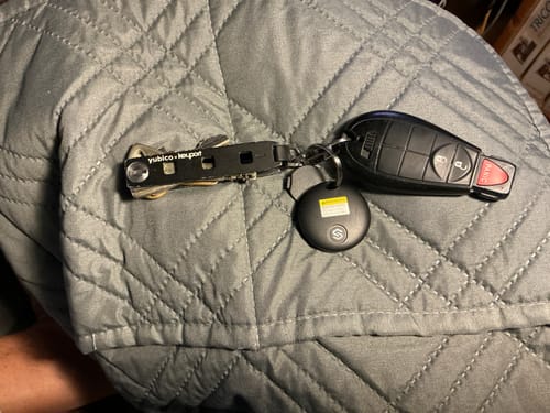 Customer's black SmartTag on a keyring with a car key fob, resting on a gray quilted surface.
