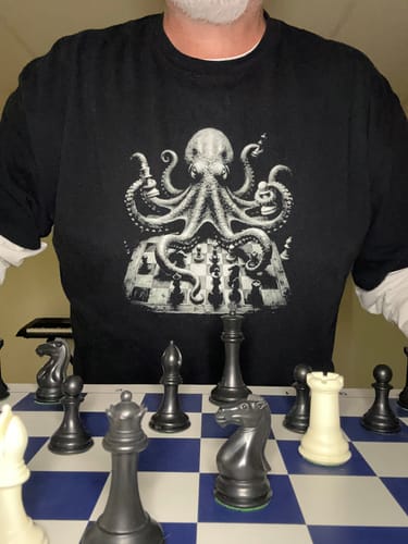 Customer wearing the black Octopus Knight t-shirt with its octopus-chess design, posed behind a real chessboard.