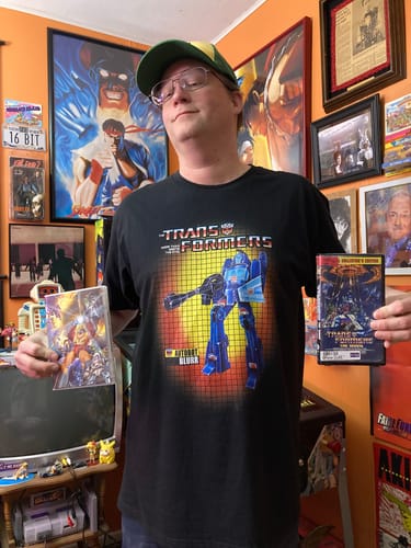 Reviewer wearing the black Box Art Blurr Transformers T-Shirt while holding other Transformers items in a decorated room.