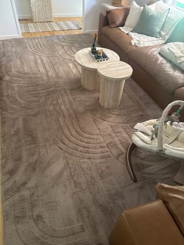 Customer's plush Mellow Mat in mocha with a carved pattern, covering a living room floor next to a leather sofa and baby bouncer.