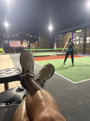 Customer relaxing in white CUURT Muuv court shoes, with an indoor court game in the background.
