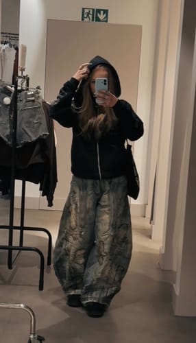 Reviewer in a fitting room wearing baggy Personsoul Stain Embroidery Curved Jeans with a black hoodie.