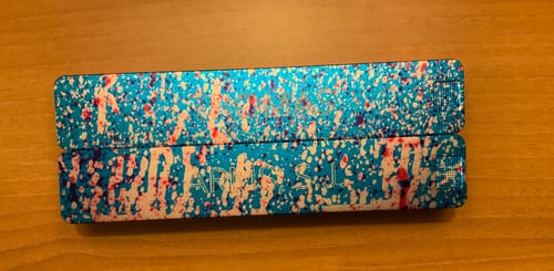 Customer's stacked Weekly AM-PM Vitamin Case with a colorful splatter-print design, lying on a wooden surface.