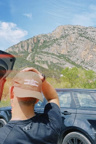 Reviewer seen from behind wearing the brown LA GORRA hat with a hand on it, outdoors with a mountain in the background.