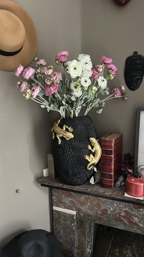 Het Ranonkel Arrangement van een klant met roze en witte bloemen in een decoratieve zwarte vaas op een schouw.