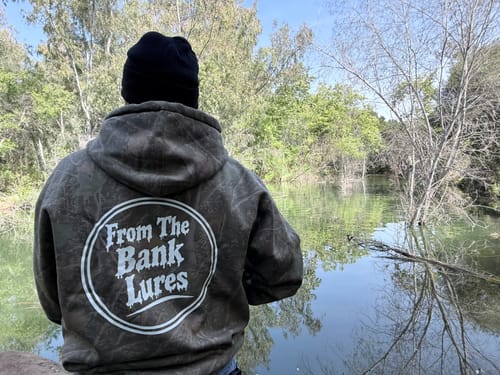 Reviewer from behind wearing the custom Camo-Styled Hoodie with a white circular logo by a river.