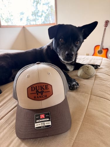 Reviewer's Custom Leather Patch Trucker Hat with 'Duke Ranch' patch on a bed, with a dog lying behind it.