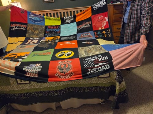 Two people holding up a large Project Repat T-Shirt Quilt made from various colorful t-shirt squares over a bed.