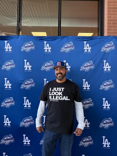 Reviewer standing while wearing the black 'I Just Look Illegal' T-shirt over a white long-sleeved shirt.