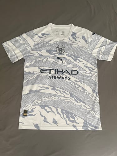 Photo d'un client du maillot Manchester City Dragon 24/25 posé à plat, montrant le motif gris sur le maillot blanc.