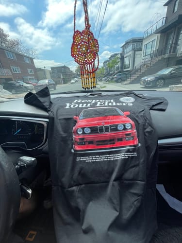 Customer's black MYSTERY TEE with a red car graphic and 'Respect Your Elders' text, displayed on a car dashboard.