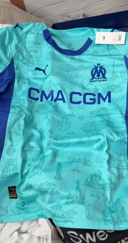 Client présentant le Maillot Spécial Marseille 25/26 déplié. Il est turquoise avec des détails et logos bleu foncé.