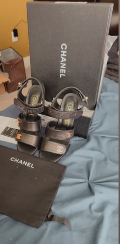 Customer's black three-strap high heel sandals shown on a bed with their original box and dust bag.