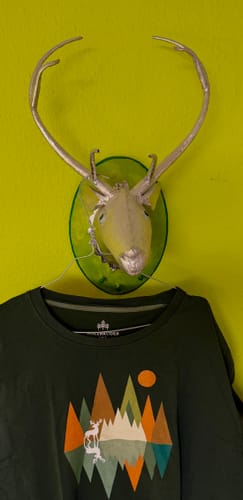 Kundenfoto des dunkelgrünen Holzbrüder-Shirts mit geometrischem Hirschmotiv, das an einer Wanddekoration hängt.