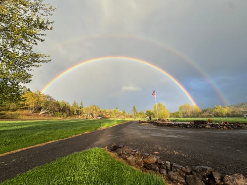 Customer's American flag on a distant flagpole, seen under a wide double rainbow spanning across a field.