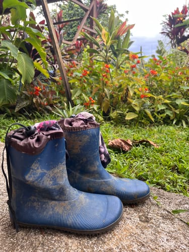 Schmutzige blaue Brisk Ⅰ Gummistiefel eines Kunden vor einer grünen Dschungelkulisse.