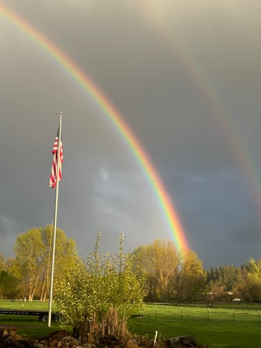 Reviewer's American flag on a flagpole with a vibrant double rainbow arching behind it against a cloudy sky.