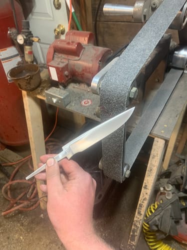 Reviewer holding a knife blade with a satin finish, with the non-woven sanding belt on a grinder in the background.