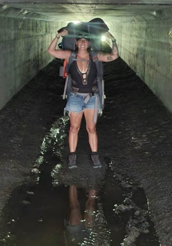 Reviewer wearing a backpack and shorts stands energetically in a dark, concrete tunnel.