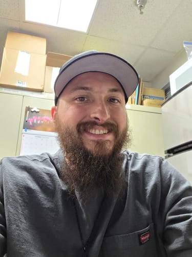 Reviewer smiling while wearing a dark gray Be The Light Hat with a light gray bill indoors.
