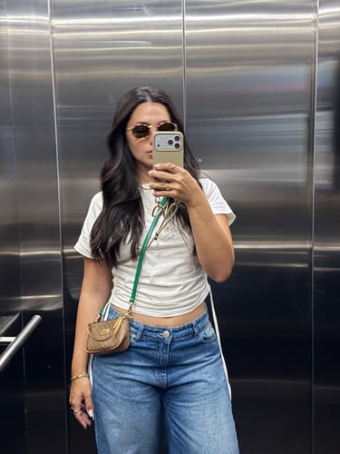 Cliente tirando uma selfie no espelho, usando seu celular com a case da coleção Nora e alça verde com detalhes dourados.